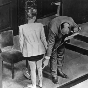 A man in a suit points to long scars on the back of a woman's calf. The picture is in black and white.